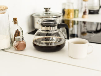 On the counter in a kitchen, a close up view of coffee maker, a cup and glass jars near a stove.