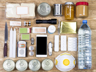Overhead view of a wooden table with items useful for an emergency including a water bottle, candles, batteries and canned food.