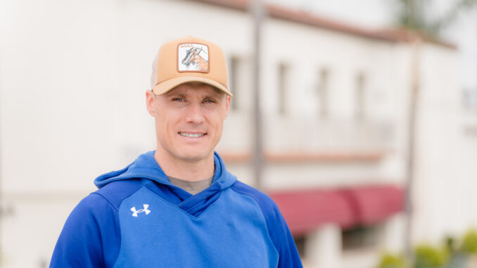 A coach stands in front of a building in a blue jacket.