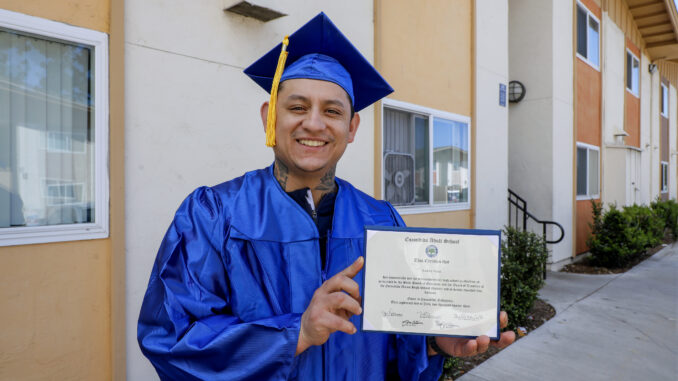 Andres Arias stands in front of his school wearing a blue graduation cap and gown and holds up his diploma.
