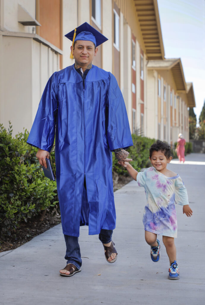 Andres Arias on the left in a blue graduation cap and gown walks down the sidewalk while holding the hand of his three year old son on the right, who is wearing an oversized tie dye shirt.
