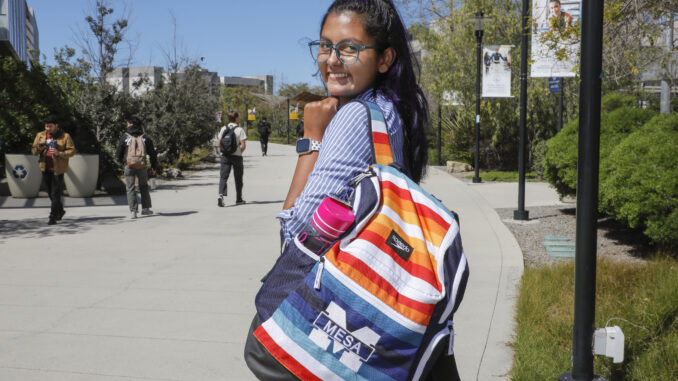 Maria Teresa Rivera stands at her college campus, her hair long and brown, wearing glasses and carrying a colorful bookbag. She is looking over her shoulder towards the camera.