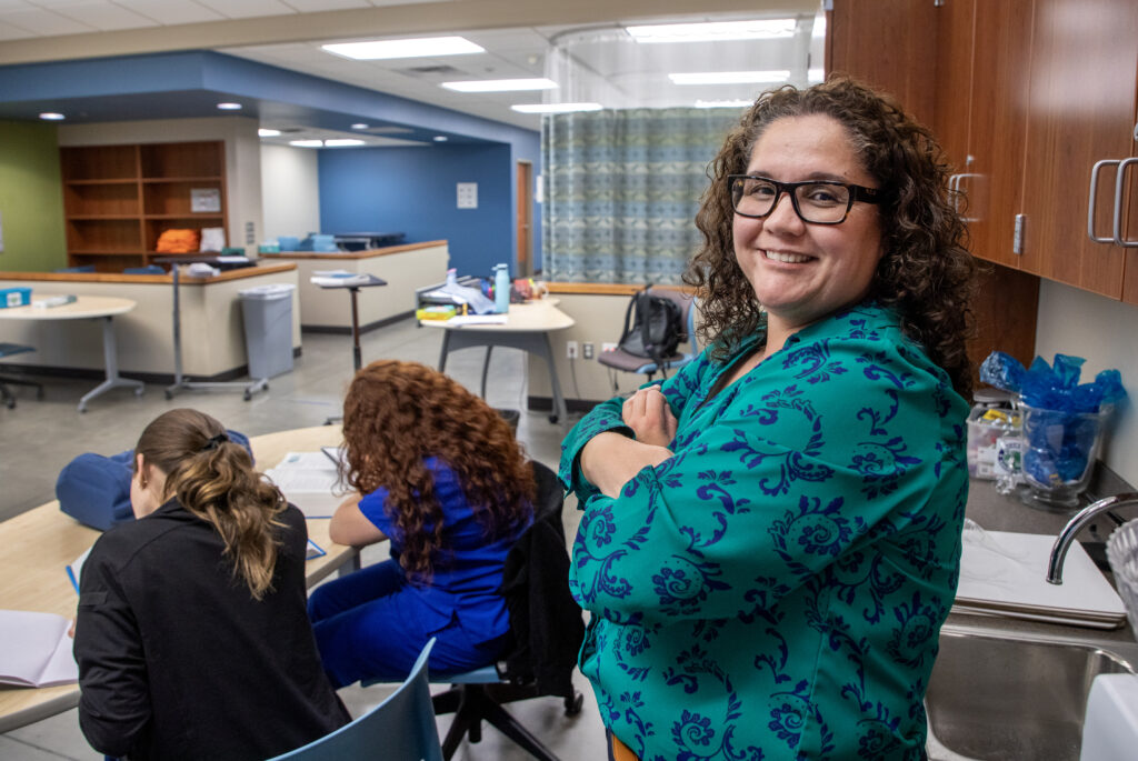 Maricxa Zendejas, a woman with dark curly hair, black rimmed glasses, and a teal blouse with a blue paisley pattern, turns and smiles for the camera in her classroom.