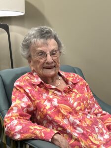 Rose Fink, and elderly woman with short gray hair and a floral blouse, sits in an armchair and smiles for the camera