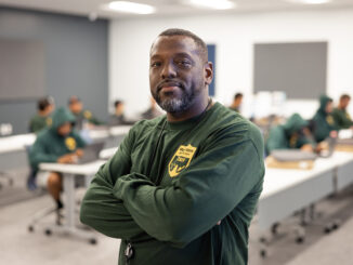 Jerry Irvin, a black man in a green long sleeved shirt, stands at his place of work with his arms crossed over his chest looking towards the camera