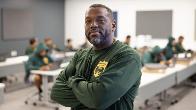 Jerry Irvin, a black man in a green long sleeved shirt, stands at his place of work with his arms crossed over his chest looking towards the camera