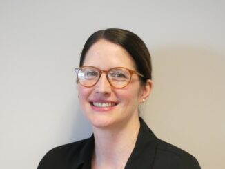 A headshot portrait of Jacqueline Hayes, a woman in a black shirt and red-rimmed glasses with her dark hair pulled back.