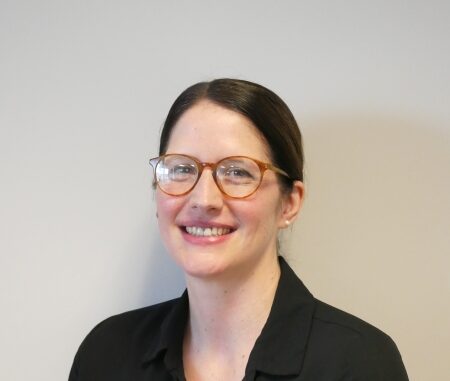 A headshot portrait of Jacqueline Hayes, a woman in a black shirt and red-rimmed glasses with her dark hair pulled back.