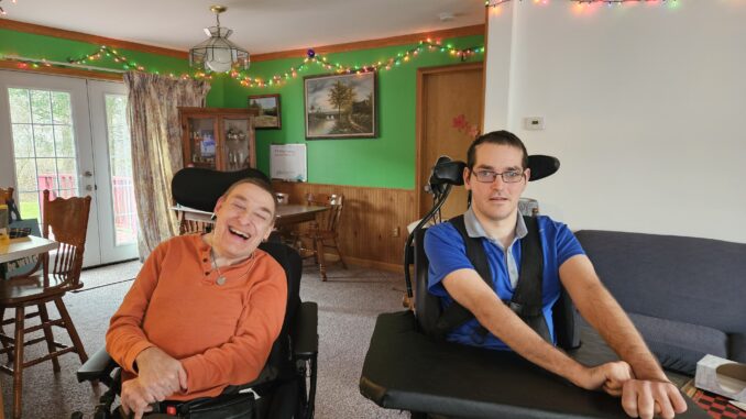 A photo of John Thompson on the left and Nick Mykytyn on right, both in motorized wheelchairs, at home with Christmas decorations in the background.