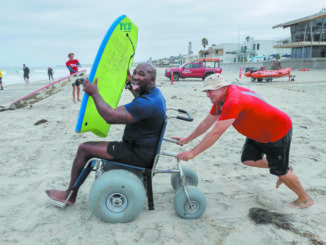 Nico Marcolongo, at right, senior manager, Operation Rebound (a program of Challenged Athletes Foundation), pushes org. member Anthony Pone, amputee Army veteran, to the water in a beach wheelchair during one of the group’s regular Wednesday morning surf sessions.