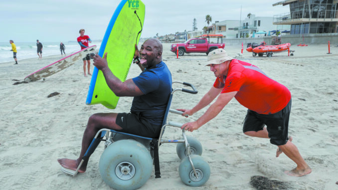 Nico Marcolongo, at right, senior manager, Operation Rebound (a program of Challenged Athletes Foundation), pushes org. member Anthony Pone, amputee Army veteran, to the water in a beach wheelchair during one of the group’s regular Wednesday morning surf sessions.