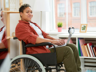 Portrait of African young woman with disabilities sitting in wheelchair and looking at camera while using laptop computer at home