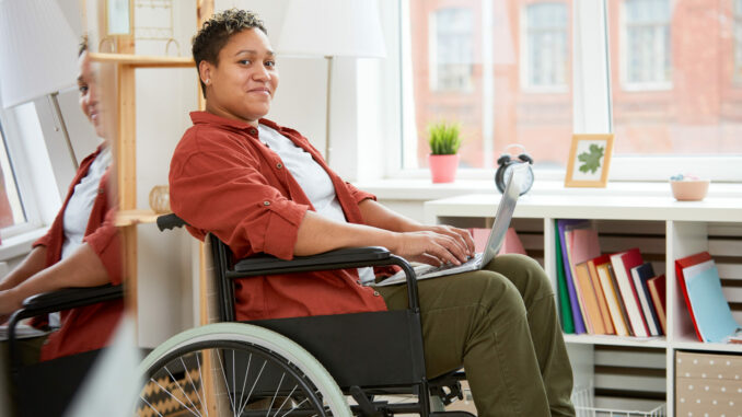 Portrait of African young woman with disabilities sitting in wheelchair and looking at camera while using laptop computer at home