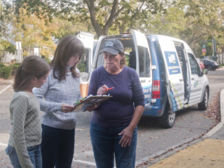 A group of people all looking at a clipboard while standing in front of a Meals on Wheels delivery van.