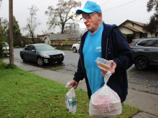 A man with a Meals on Wheels shirt and hat carrying bagged food on a front lawn.