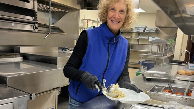 A woman, working behind a professional kitchen counter, serving up some food on a plate.