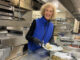 A woman, working behind a professional kitchen counter, serving up some food on a plate.