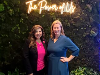 Two women standing next to each other smiling with a neon sign in the background that says, "the power of us".