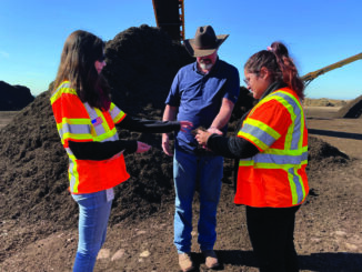 Three people gathered in front of a large compost pile. One person is holding a hand full of dirt and all three people are inspecting it.