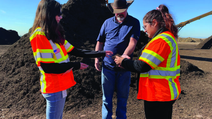 Three people gathered in front of a large compost pile. One person is holding a hand full of dirt and all three people are inspecting it.