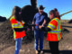 Three people gathered in front of a large compost pile. One person is holding a hand full of dirt and all three people are inspecting it.