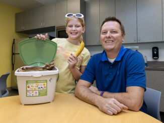 Father and daughter sitting at table with a countertop organic waste pail. The daughter is holding a banana.