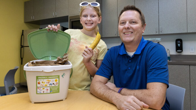 Father and daughter sitting at table with a countertop organic waste pail. The daughter is holding a banana.
