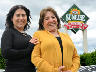 Two women standing next to each other smiling with a sign for the Sunrise adult day care center in the background.