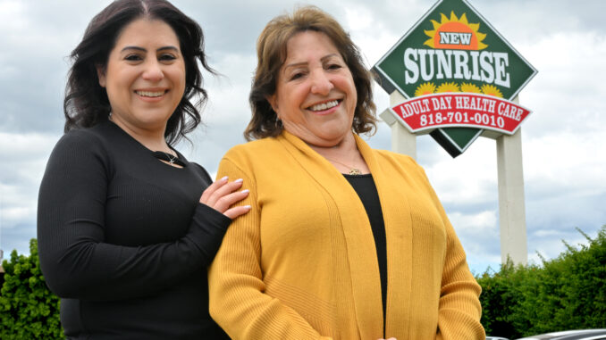 Two women standing next to each other smiling with a sign for the Sunrise adult day care center in the background.