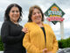 Two women standing next to each other smiling with a sign for the Sunrise adult day care center in the background.