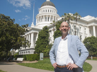 A man, standing with his hands on his hips smiling. He is standing in front of the California state capitol.