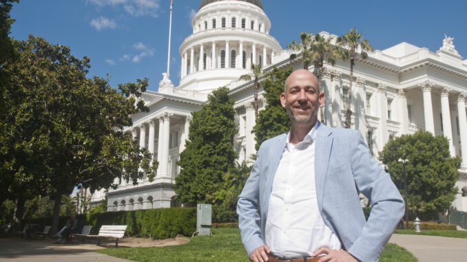 A man, standing with his hands on his hips smiling. He is standing in front of the California state capitol.