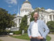 A man, standing with his hands on his hips smiling. He is standing in front of the California state capitol.